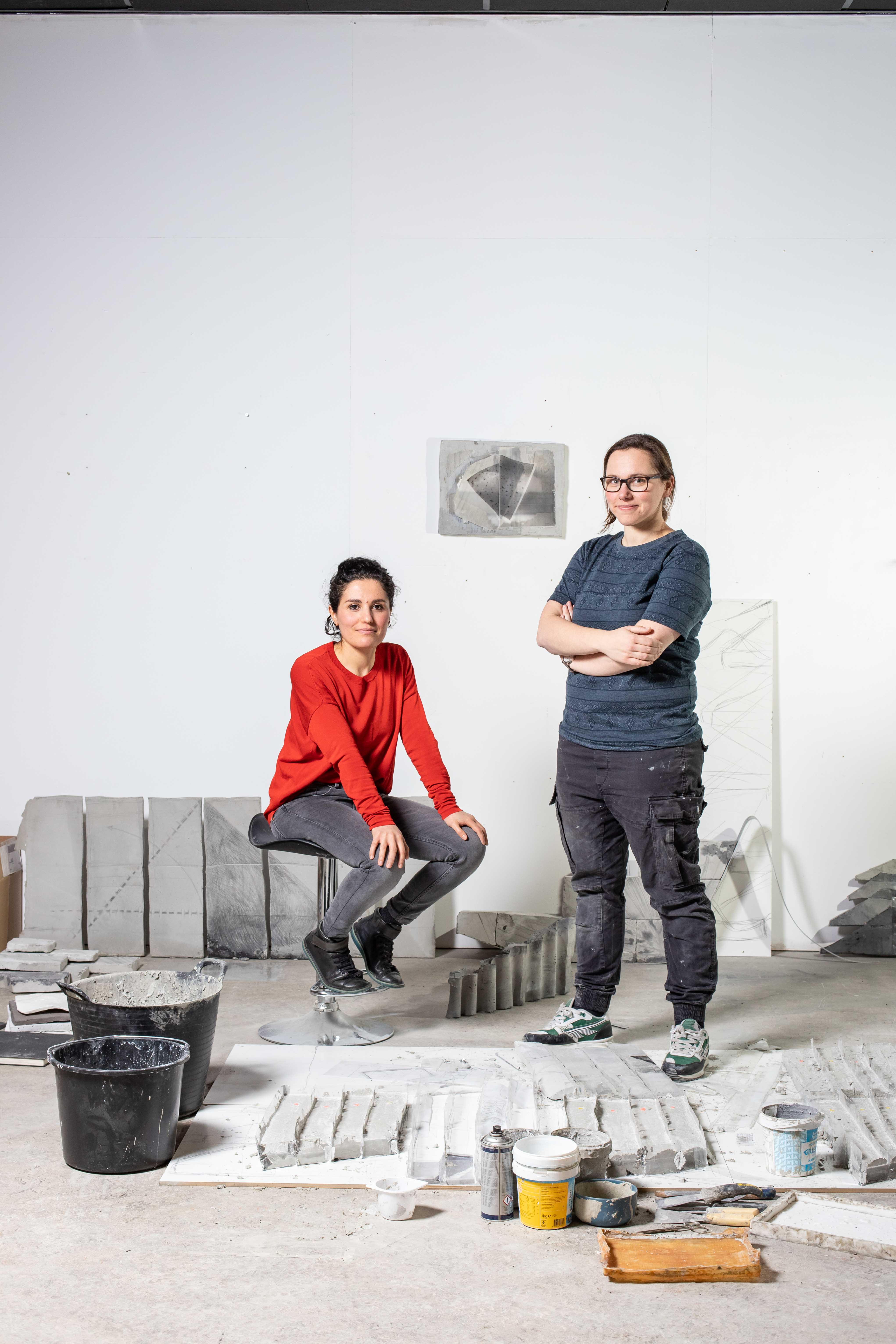 Natalia Ossef zittend op een krukje en Maaike Kramer staand in het atelier van Maaike met betonnen objecten en gereedschap op de vloer rondom hen